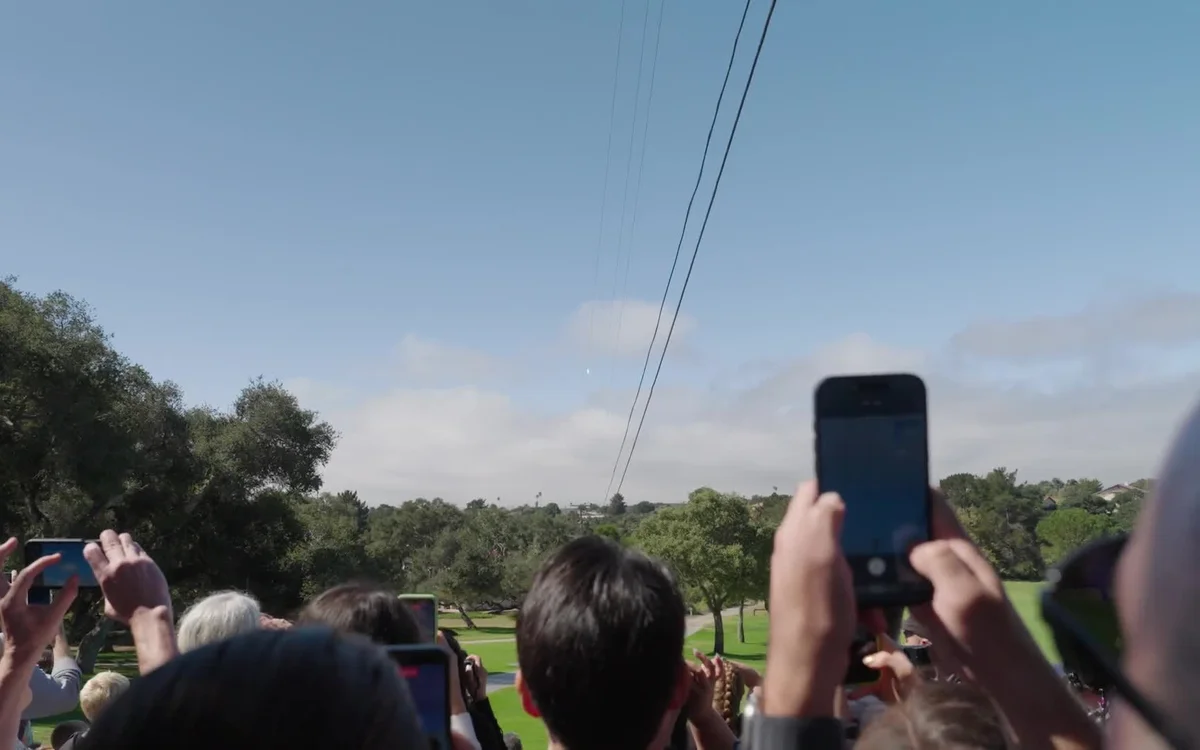 A crowd outdoors holding up phones to film a Falcon 9 rocket climbing on a thin exhaust trail through a blue sky